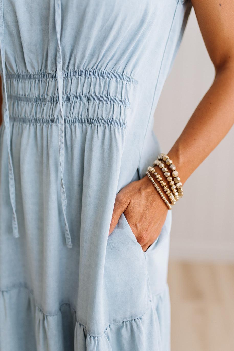 Close-up of a person wearing a light blue smocked dress, with one hand in a side pocket and stacked gold beaded bracelets on the wrist.