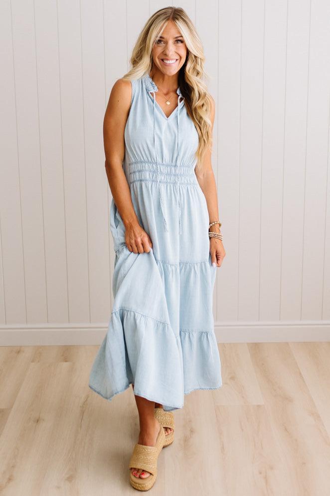 A woman modeling a sleeveless light blue tiered maxi dress with a smocked waist, standing on a light wood floor against a white paneled wall.