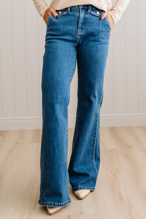 WOMAN WEARING HIGH RISE FLARE JEANS IN A DARK WASH AND BUTTON FLAP SMALL POCKETS AT THE HIP.