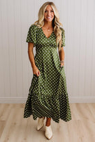 A woman modeling a green midi wrap dress with white polka dots, featuring short flutter sleeves, a V-neckline, and a tiered ruffle hem, paired with cream ankle boots against a white paneled wall and light wood floor.