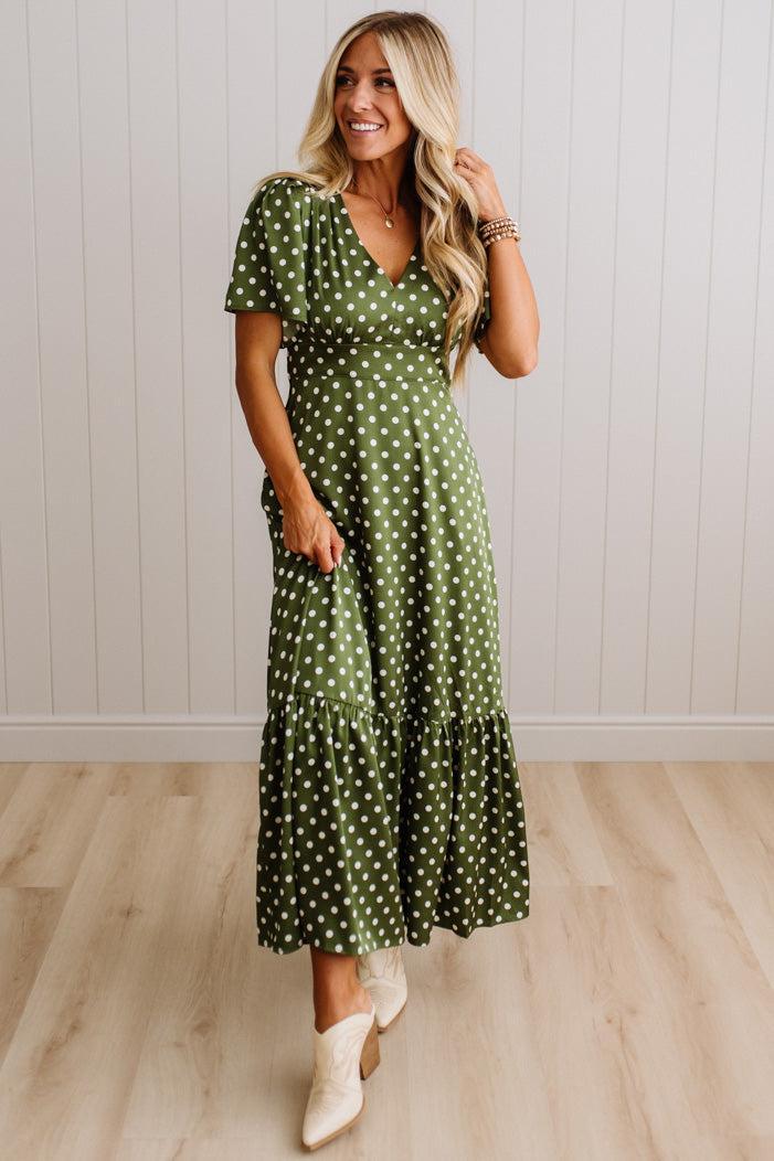 A woman modeling a green midi dress with white polka dots, featuring short puff sleeves, a V-neckline, and a tiered ruffle hem, paired with white cowboy boots against a white paneled wall and light wood floor.