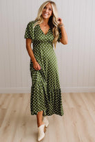 A woman modeling a green midi dress with white polka dots, featuring short flutter sleeves, a V-neckline, and a tiered skirt, paired with white cowboy boots against a light wood-paneled wall.