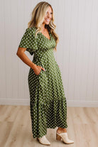 A woman modeling a green and white polka dot maxi dress with short flutter sleeves, a V-neckline, and side pockets, paired with cream-colored heeled mule boots, set against a white paneled wall and light wood floor.