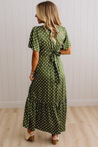 A woman wearing a green and white polka dot midi dress with short puff sleeves and a tie waist, shown from the back against a white paneled wall and light wood floor.