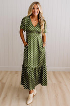 A woman modeling a green short-sleeve maxi dress with white polka dots, standing in front of a white paneled wall and light wood floor.