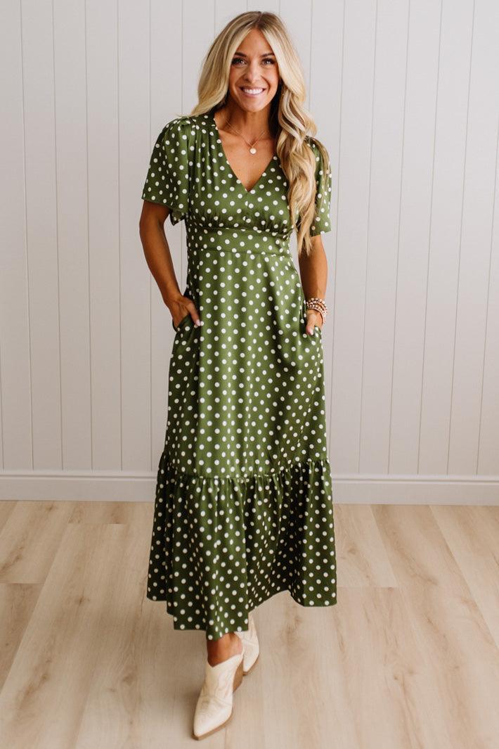 A woman modeling a green short-sleeve maxi dress with white polka dots, standing in front of a white paneled wall and light wood floor.