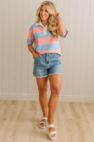 Woman wearing a colorful striped shirt and denim shorts standing against a light wooden paneled wall.