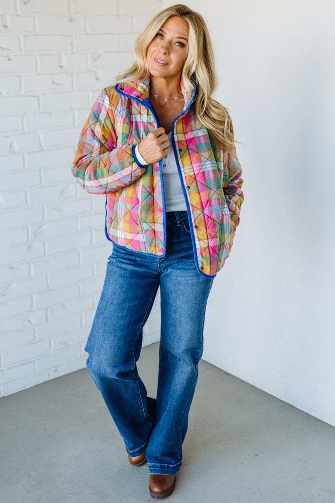 Woman wearing a colorful plaid jacket and blue jeans against a white brick wall.