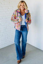 Woman wearing a colorful plaid jacket, white shirt, blue jeans, and brown shoes against a white brick wall.
