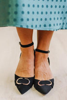 WOMAN IN A GREEN POLKA DOT DRESS WITH BLACK LOW HEELS, POINTED TOE , ANKLE STRAP AND GOLD BUCKLE ACCENT.