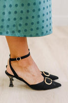 WOMAN IN A GREEN POLKA DOT DRESS WITH BLACK LOW HEELS, POINTED TOE , ANKLE STRAP AND GOLD BUCKLE ACCENT.