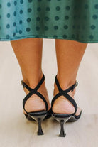 WOMAN IN A GREEN POLKA DOT DRESS WITH BLACK LOW HEELS, POINTED TOE , ANKLE STRAP AND GOLD BUCKLE ACCENT.