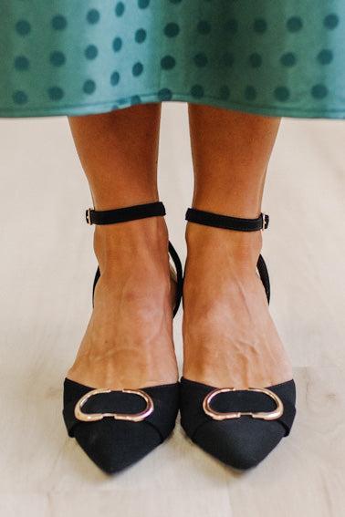 WOMAN IN A GREEN POLKA DOT DRESS WITH BLACK LOW HEELS, POINTED TOE , ANKLE STRAP AND GOLD BUCKLE ACCENT.