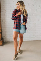 Woman wearing a red plaid shirt, white top, and denim shorts against a white brick wall.
