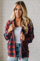 Woman wearing a red and blue plaid shirt over a white top and denim jeans against a white brick wall.