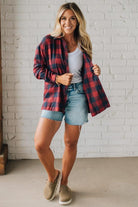 Woman wearing a red plaid shirt, white tank top, and denim shorts against a white brick wall.