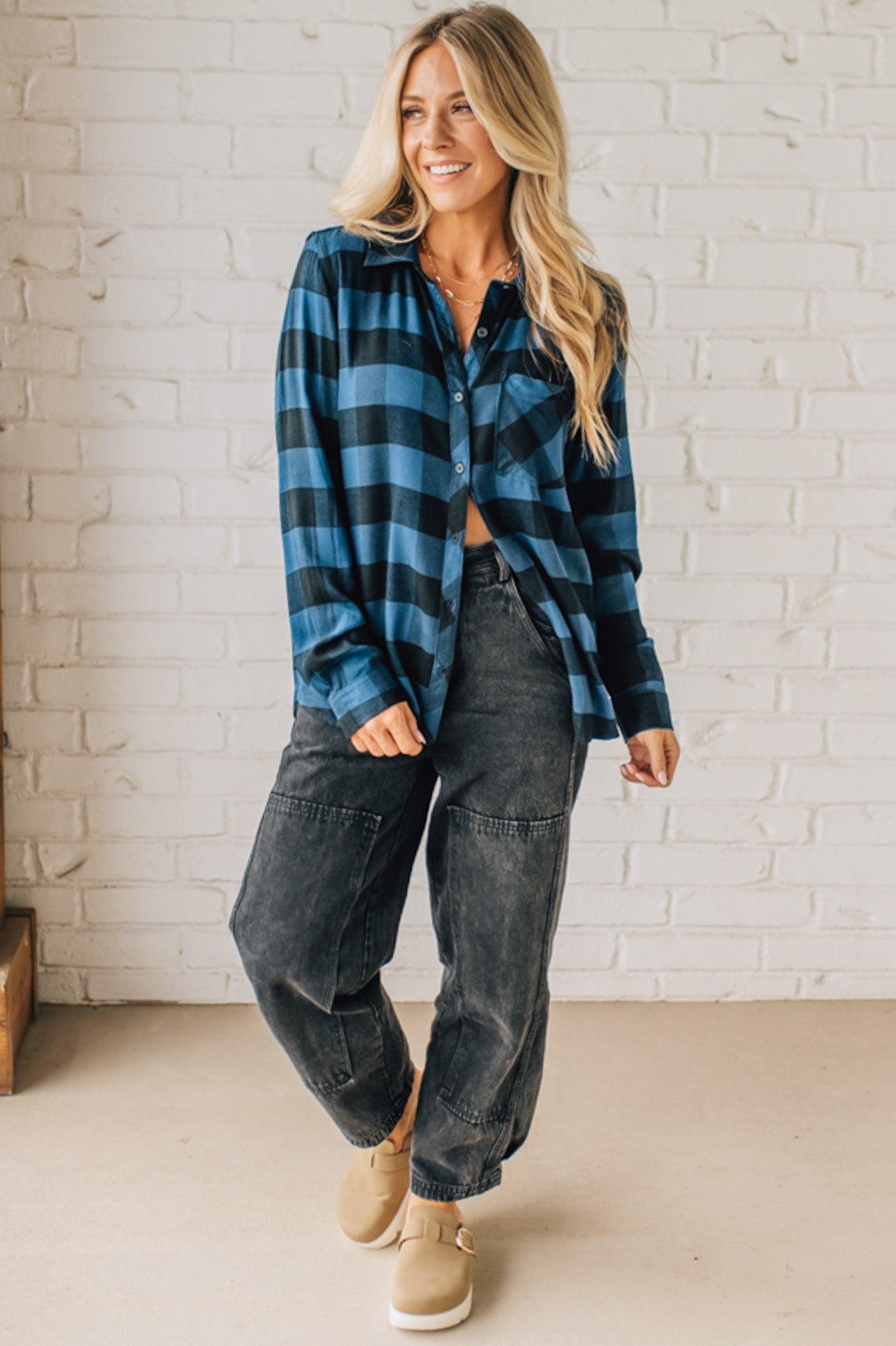 Woman wearing a blue plaid shirt and dark jeans standing against a white brick wall.