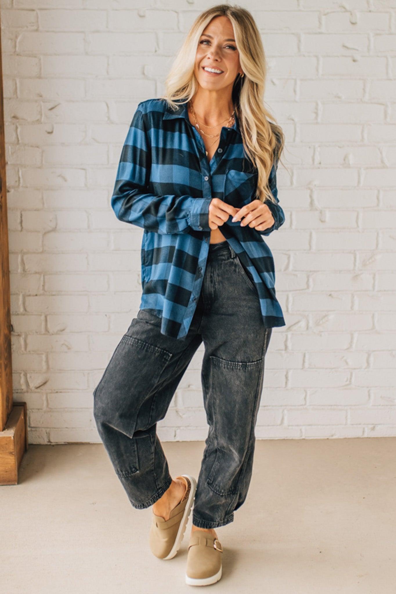 Woman wearing a blue plaid shirt and dark jeans standing against a white brick wall.