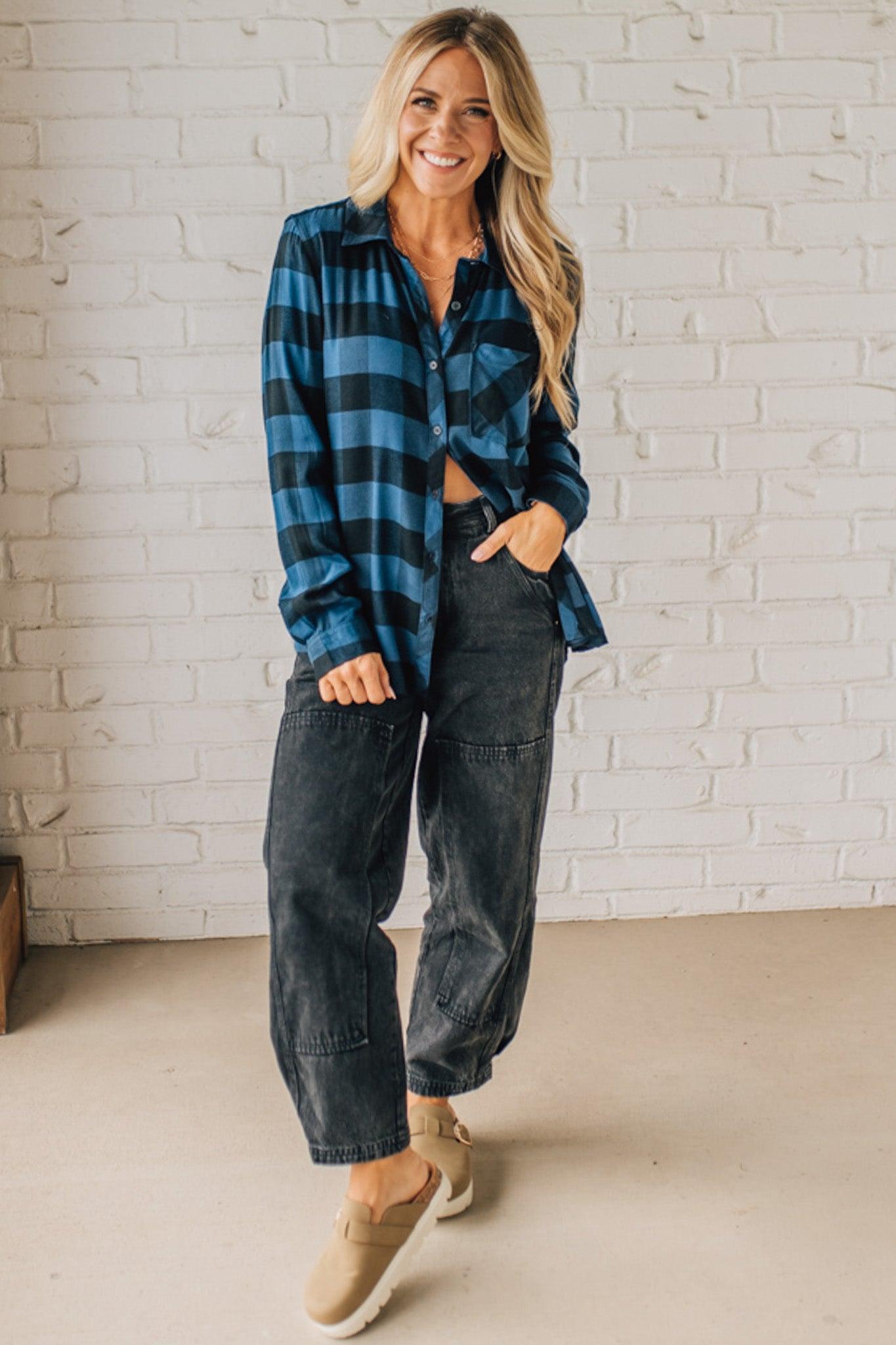 Woman wearing a blue and black plaid shirt and dark jeans against a white brick wall.
