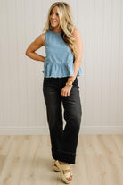 Woman wearing a light blue sleeveless top and black pants standing against a white paneled wall.