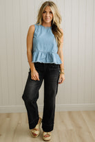 Woman wearing a light blue sleeveless top and black jeans against a white paneled wall.