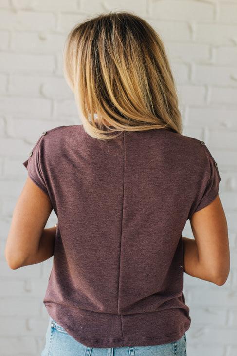 Blonde woman wearing a heathered deep berry color tee shirt with two straps on the shoulder with a button on each end of the strap.