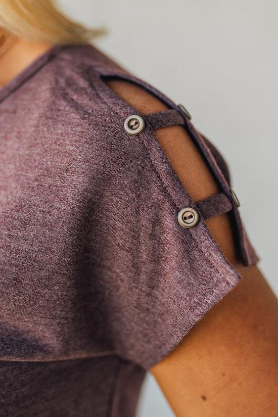 Blonde woman wearing a heathered deep berry color tee shirt with two straps on the shoulder with a button on each end of the strap.