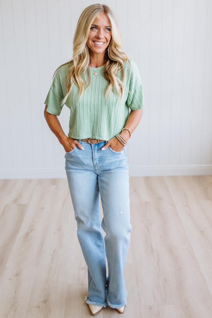 A woman stands in a bright room with light wood floors and white paneled walls, wearing a light sage green short-sleeve ribbed knit top, light-wash blue jeans, and beige pointed-toe heels.