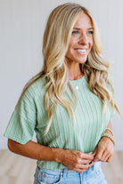 A smiling blonde woman wearing a light mint green short-sleeve knit top with a round neckline and light blue jeans, photographed against a soft light gray indoor background.