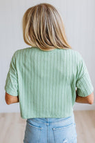 A woman viewed from behind wearing a mint green short-sleeve ribbed knit top and light blue jeans, standing against a plain light gray wall.