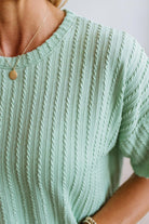 Close-up of a person wearing a mint green short-sleeve ribbed knit top with vertical cable-like texture and a delicate gold pendant necklace.