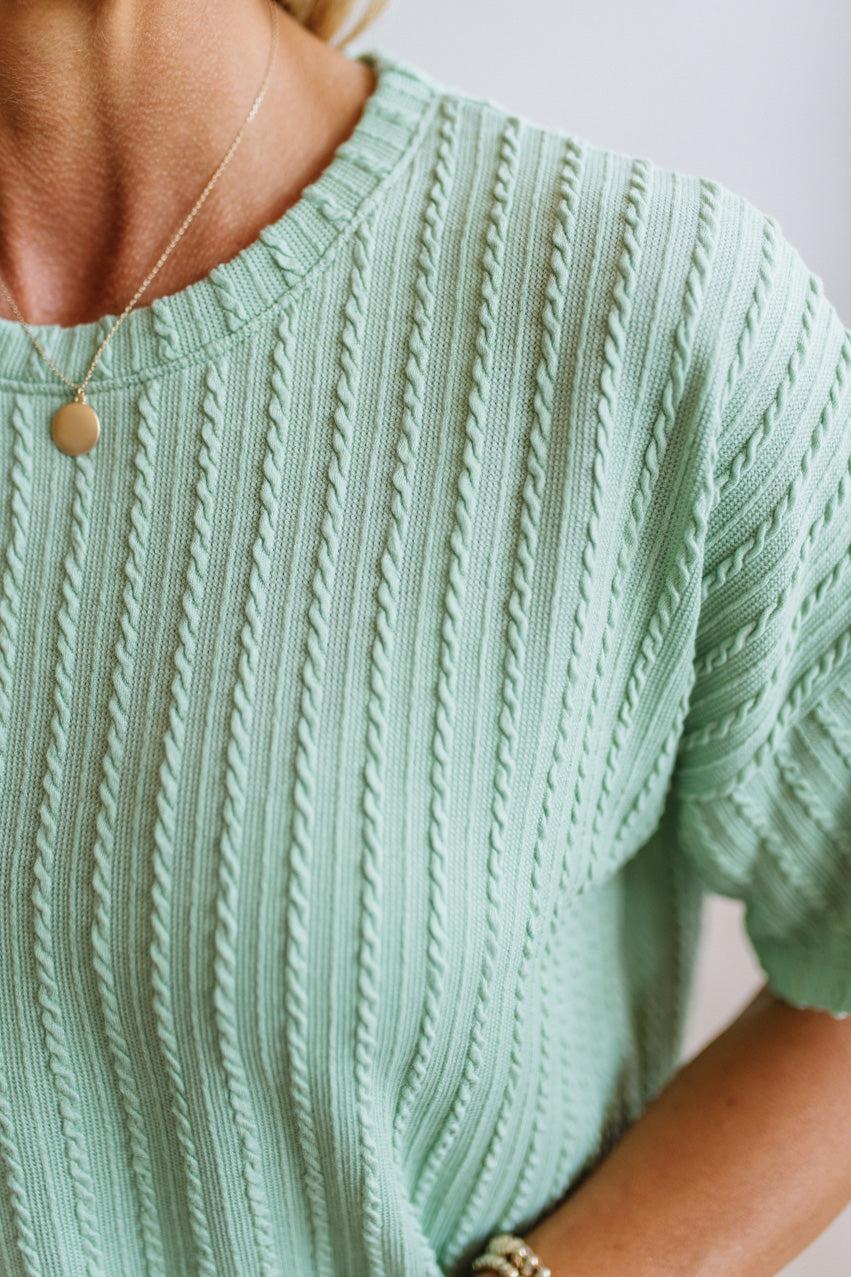 Close-up of a person wearing a mint green short-sleeve ribbed knit top with vertical cable-like texture and a delicate gold pendant necklace.