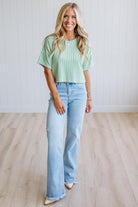 A woman modeling a light mint green short-sleeve ribbed top and light-wash blue flare jeans, standing on a light wood floor against a white paneled wall.