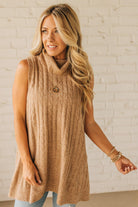 Woman wearing a beige sleeveless sweater against a white brick wall.