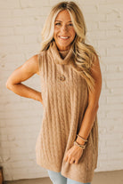 Woman wearing a beige sleeveless sweater over a yellow top against a white brick wall.