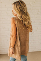 Woman wearing a brown sleeveless sweater against a white brick wall.