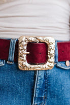 A rippled square frame buckle with a molten, hammered gold-tone finish sits front and center, on a burgundy faux suede belt.