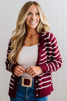 BLONDE WOMAN IN A BRIGHTLY LIT STUDIO WEARING A CREAM TANK AND DARK JEANS SHOWING A OUTSEAMED, LONG SLEEVE, FUZZY, CREW NECK STRIPED BUTTON DOWN SWEATER CARDIGAN IN BURGUNDY.