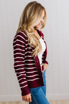 BLONDE WOMAN IN A BRIGHTLY LIT STUDIO WEARING A CREAM TANK AND DARK JEANS SHOWING A OUTSEAMED, LONG SLEEVE, FUZZY, CREW NECK STRIPED BUTTON DOWN SWEATER CARDIGAN IN BURGUNDY.