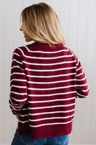 BLONDE WOMAN IN A BRIGHTLY LIT STUDIO WEARING A CREAM TANK AND DARK JEANS SHOWING A OUTSEAMED, LONG SLEEVE, FUZZY, CREW NECK STRIPED BUTTON DOWN SWEATER CARDIGAN IN BURGUNDY.