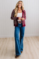 BLONDE WOMAN IN A BRIGHTLY LIT STUDIO WEARING A CREAM TANK AND DARK JEANS SHOWING A OUTSEAMED, LONG SLEEVE, FUZZY, CREW NECK STRIPED BUTTON DOWN SWEATER CARDIGAN IN BURGUNDY.