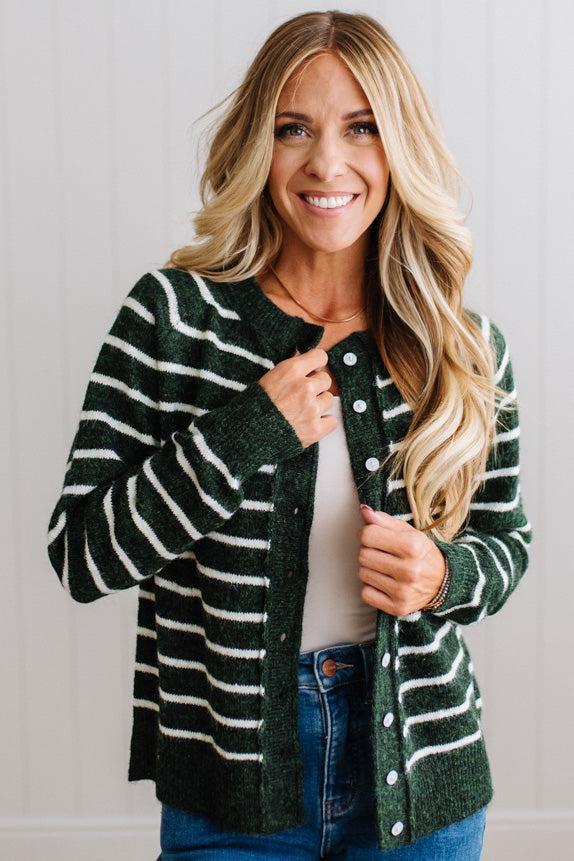 BLONDE WOMAN IN A BRIGHTLY LIT STUDIO WEARING A CREAM TANK AND DARK JEANS SHOWING A OUTSEAMED, LONG SLEEVE, FUZZY, CREW NECK STRIPED BUTTON DOWN SWEATER CARDIGAN IN FOREST GREEN.