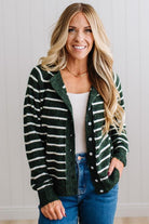 BLONDE WOMAN IN A BRIGHTLY LIT STUDIO WEARING A CREAM TANK AND DARK JEANS SHOWING A OUTSEAMED, LONG SLEEVE, FUZZY, CREW NECK STRIPED BUTTON DOWN SWEATER CARDIGAN IN FOREST GREEN.