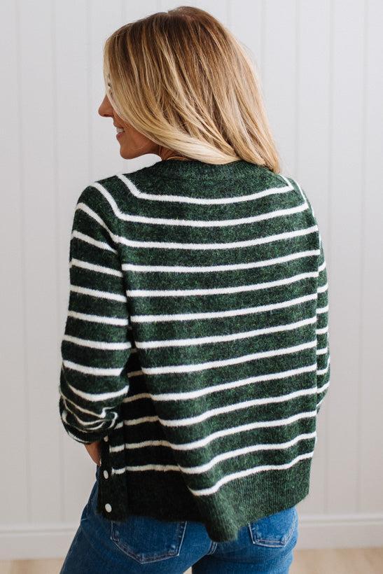 BLONDE WOMAN IN A BRIGHTLY LIT STUDIO WEARING A CREAM TANK AND DARK JEANS SHOWING A OUTSEAMED, LONG SLEEVE, FUZZY, CREW NECK STRIPED BUTTON DOWN SWEATER CARDIGAN IN FOREST GREEN.