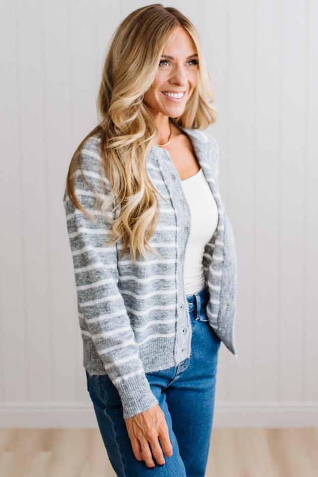 BLONDE WOMAN IN A BRIGHTLY LIT STUDIO WEARING A CREAM TANK AND DARK JEANS SHOWING A OUTSEAMED, LONG SLEEVE, FUZZY, CREW NECK STRIPED BUTTON DOWN SWEATER CARDIGAN IN HEATHER GREY.