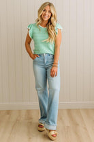 Woman wearing a green top and blue jeans standing against a light wooden paneled wall.