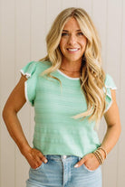 Woman wearing a light green top with ruffled sleeves and blue jeans, standing against a neutral background.