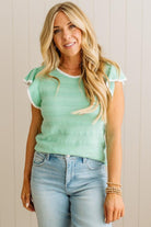Woman wearing a light green top with ruffled sleeves and blue jeans against a beige wall.
