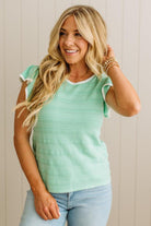 Woman wearing a light green sleeveless top and blue jeans against a beige wall.