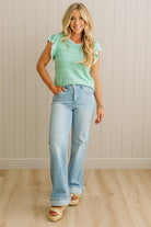 Woman wearing a light green top and blue jeans standing against a beige wall.